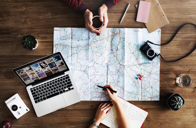 top-view photography of persons holding mug and pen using MacBook and world map, planning for travel